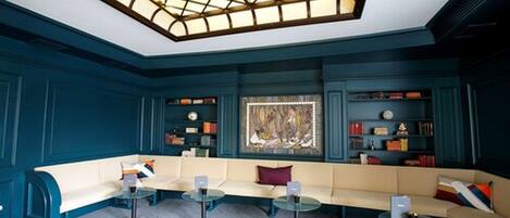 A modern lounge with a blue color scheme, featuring a large stained glass window, a sofa area with tables, and bookshelves.