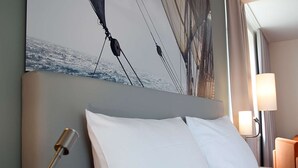 A hotel room with a large bed, white pillows, a headboard with a painting, a bedside lamp, and a window with curtains. - Nordsee Hotel Bremerhaven City (Bremerhaven)