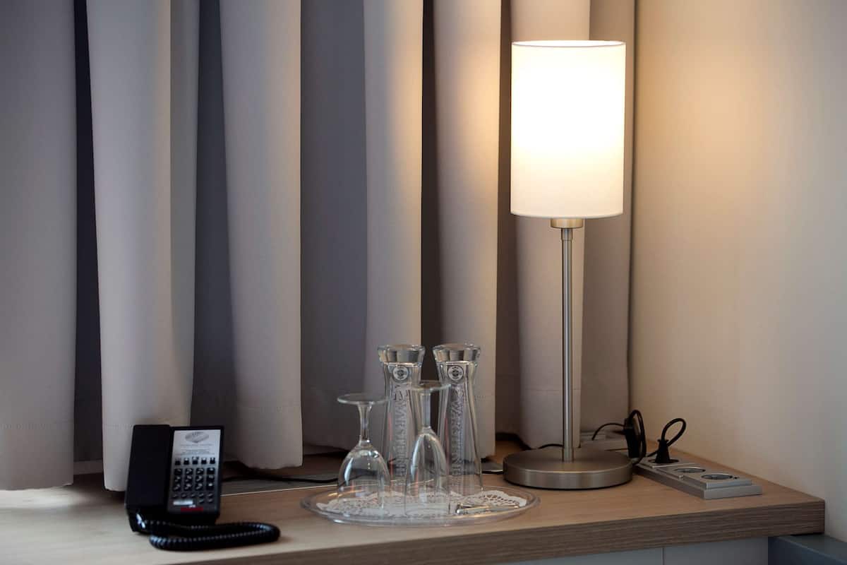 Habitación de hotel con una mesita de noche que tiene un teléfono, una lámpara y una bandeja con tres jarrones de vidrio transparente.
