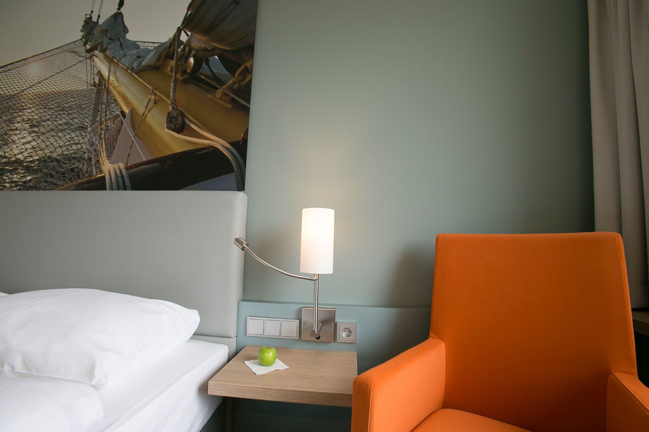 A hotel room with a bed, a nightstand with a lamp, an orange chair, and a wall-mounted TV.