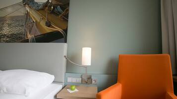Una habitación de hotel con una cama, una mesita de noche con lámpara, un sillón naranja y un televisor montado en la pared.