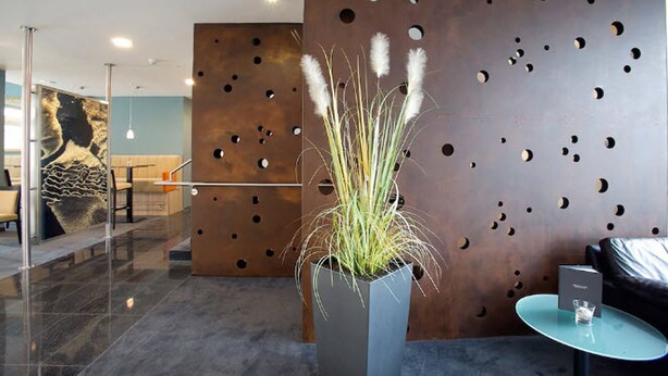 A modern lobby with a glass partition, a metal wall with circular cutouts, a potted plant, a blue table, and a sofa. - Nordsee Hotel Bremerhaven City (Bremerhaven)