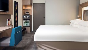 A hotel room with a bed, a desk, a chair, a television, and a minibar. - Novotel Paris Rueil Malmaison (Rueil-Malmaison)