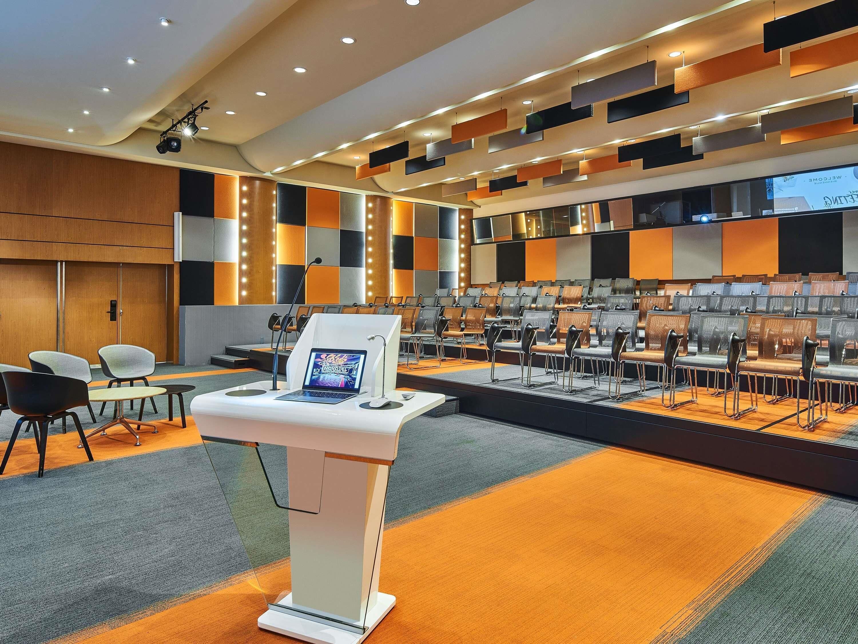 Une salle de conférence équipée d’une scène, de rangées de chaises, d’un pupitre et d’un grand écran.