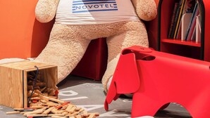 A large teddy bear in a room with orange walls, a bookshelf, and a framed picture of rainbows. - Novotel Paris Rueil Malmaison (Rueil-Malmaison)