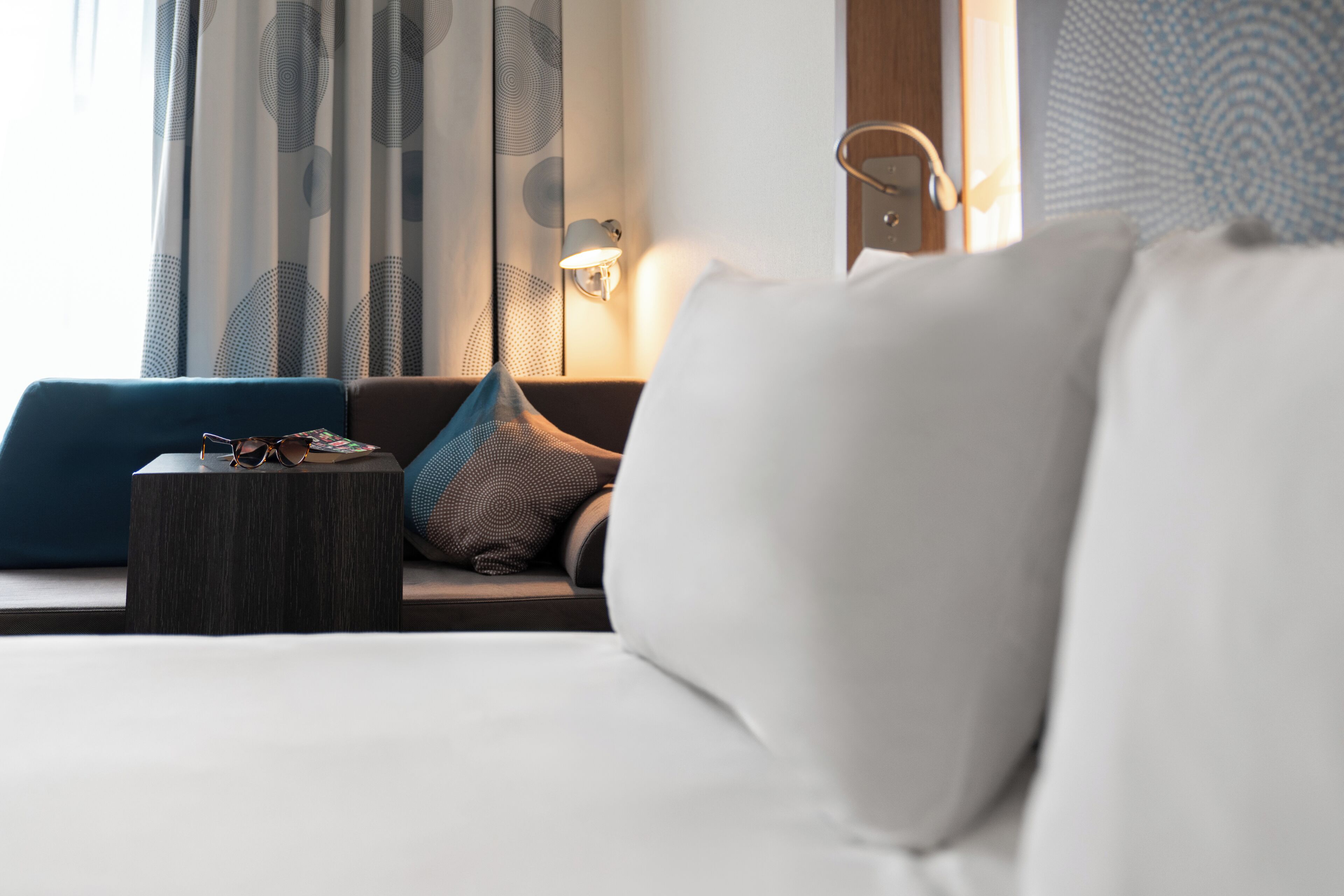 Ein Hotelzimmer mit Bett, Nachttisch, Lampe und Blick durch Vorhänge nach draußen.