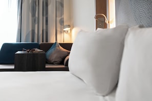 Habitación de hotel con cama, mesita auxiliar, lámpara y vistas al exterior a través de cortinas.