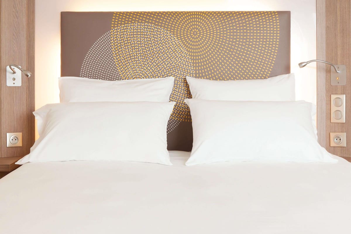 A neatly made bed with white linens, a modern abstract wall art piece above it, and a wooden headboard.