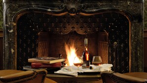 Ein Kamin mit brennendem Feuer, eine Weinflasche, Gläser und Bücher auf einem Tisch.