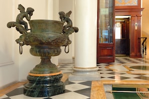 Eine prachtvolle Halle mit einer großen, verzierten Vase auf einem Marmorsockel, flankiert von hohen Säulen, und einem Schachbrettmuster auf dem Boden.