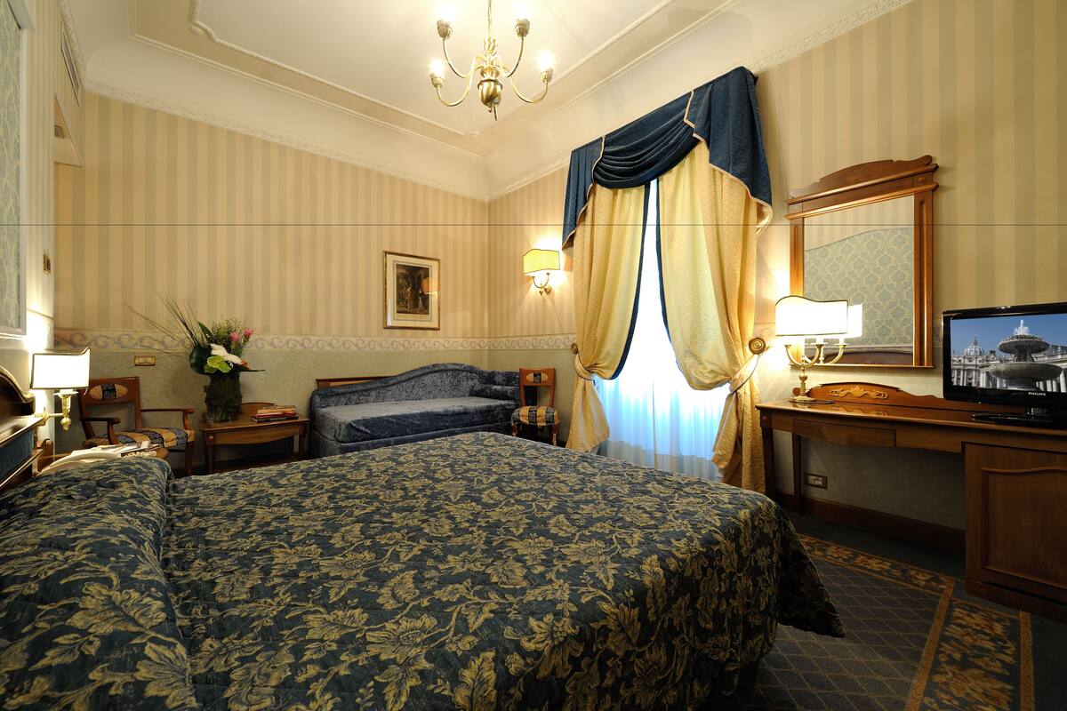 A hotel room with a large bed, a desk with a television, a chair, a nightstand, and a lamp.