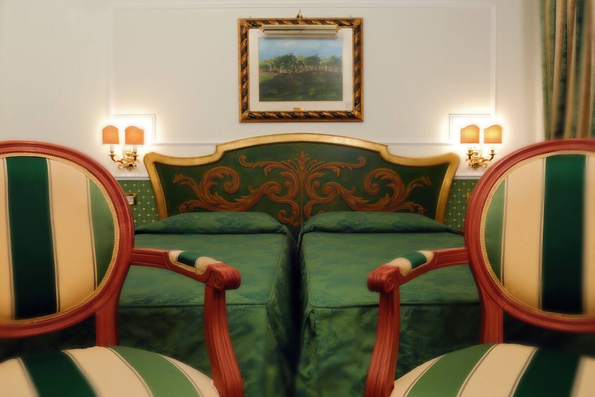 A room with a bed, two chairs, a framed picture on the wall, and wall sconces.