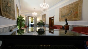 Ein Flügel in einem großzügigen Raum mit einer Chandelüster, großen Gemälden und einem roten Sofa.