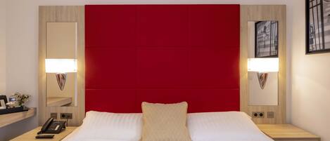Habitación de hotel con una cama grande, cabecero rojo, lámparas de noche, mesita de noche con un teléfono y un espejo.