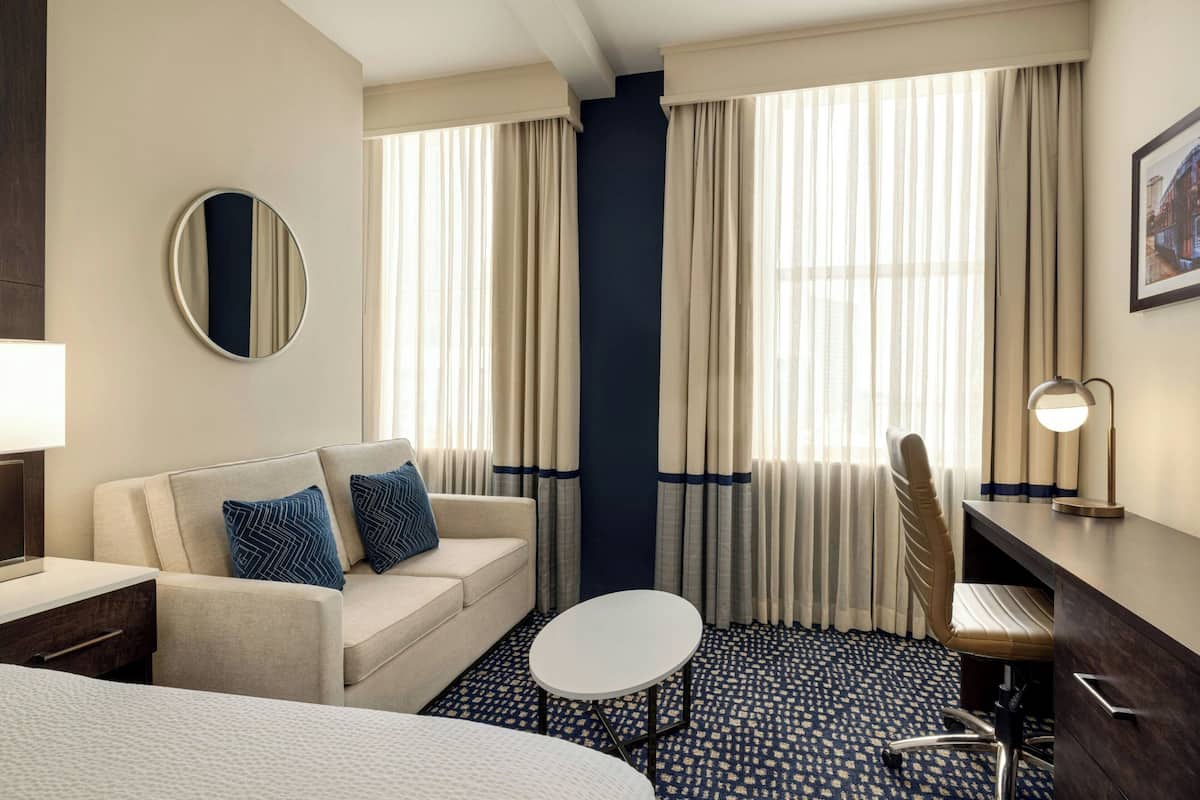 Una habitación de hotel con un sofá, un espejo redondo, un escritorio, una silla y una pequeña mesa.