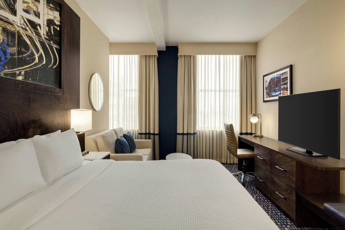 A hotel room with a large bed, a desk with a flat-screen TV, a chair, a lamp, and a large window with curtains.