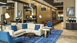 A modern hotel lobby with a seating area, a coffee station, and a large mirror reflecting the space.