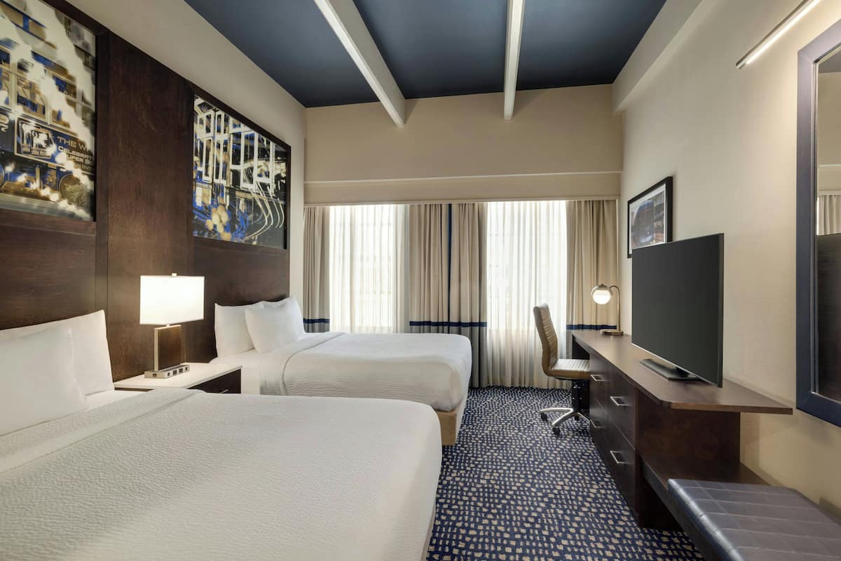Habitación de hotel con dos camas, un escritorio con televisor, una silla y un ventanal grande con cortinas.