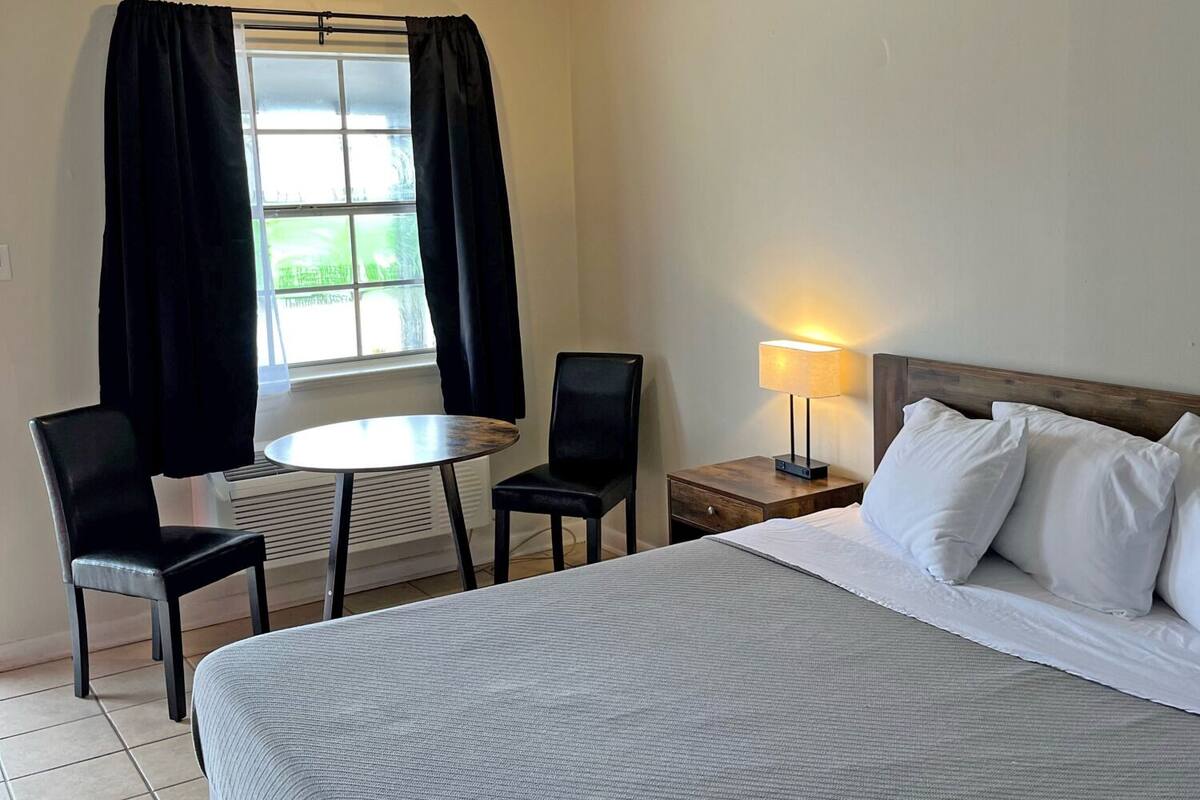 A hotel room with a bed, a table, two chairs, a lamp, and a window with curtains.