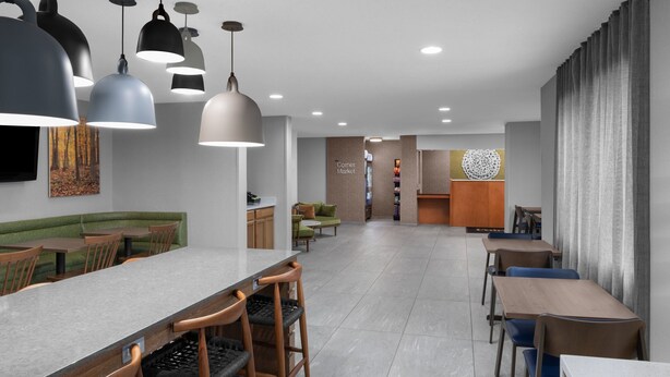 Un espace de salle à manger moderne avec une longue table, de nombreuses chaises et des luminaires suspendus. On y trouve également un coin salon avec un canapé et des fauteuils, ainsi qu’un comptoir avec des tabourets.