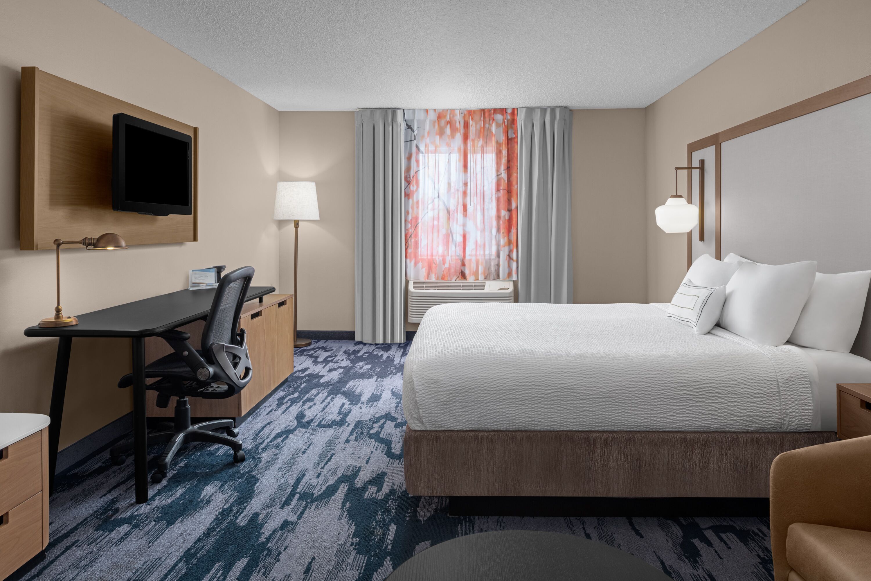 Habitación de hotel con cama, escritorio, silla, televisor, lámpara y ventana con cortinas.