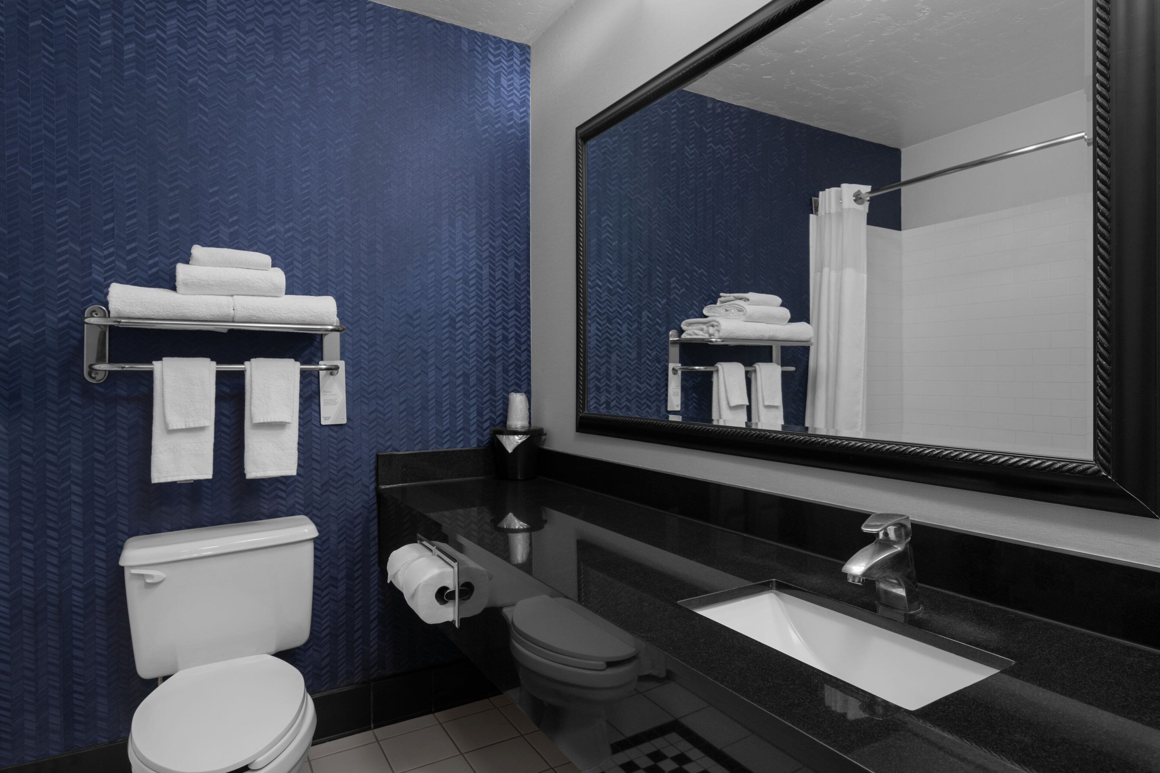 A modern bathroom with a black countertop, white sink, and toilet. There is a towel rack with folded towels and a large mirror above the sink.