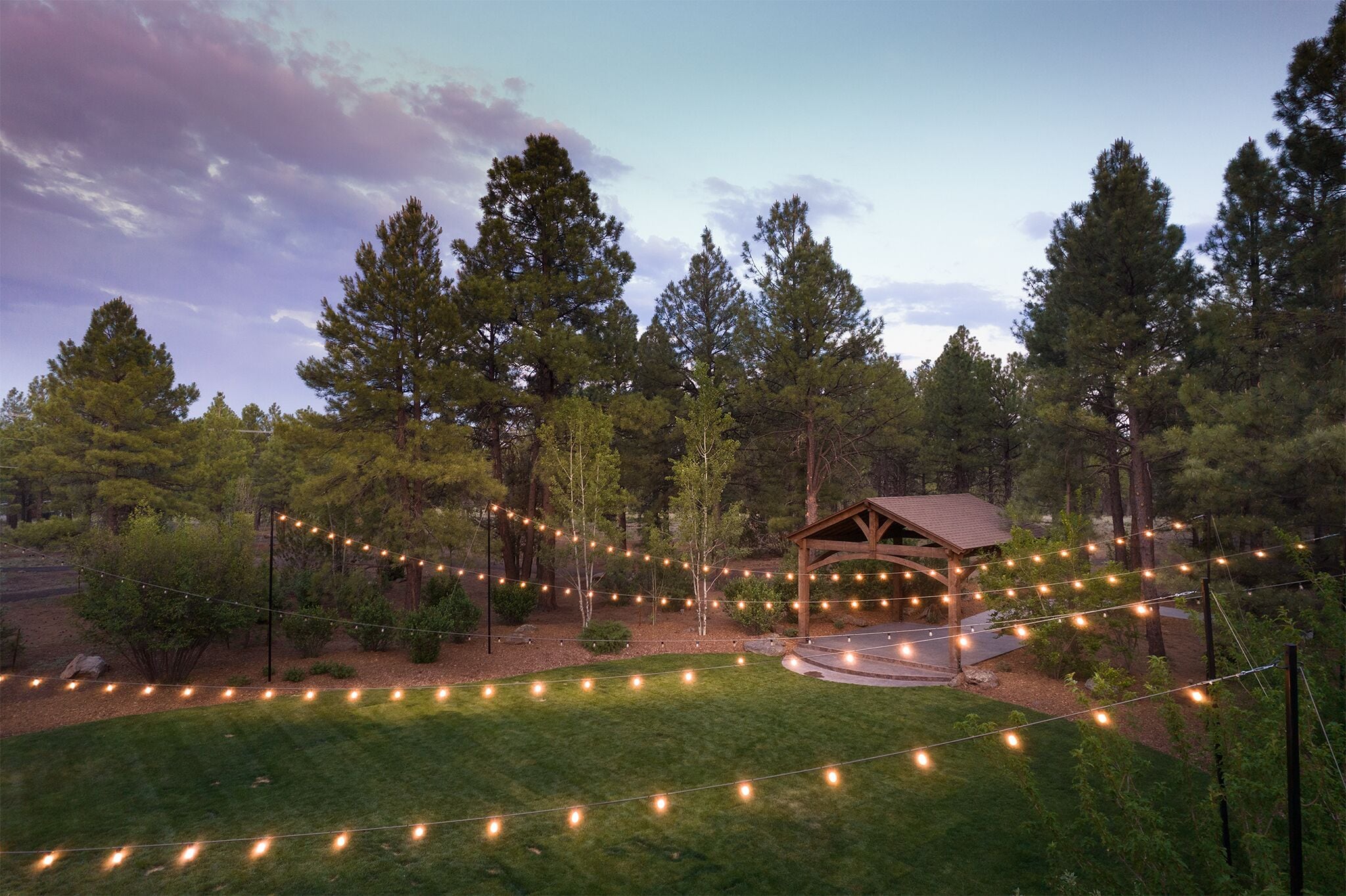A gazebo surrounded by a lawn with string lights, set in a wooded area.