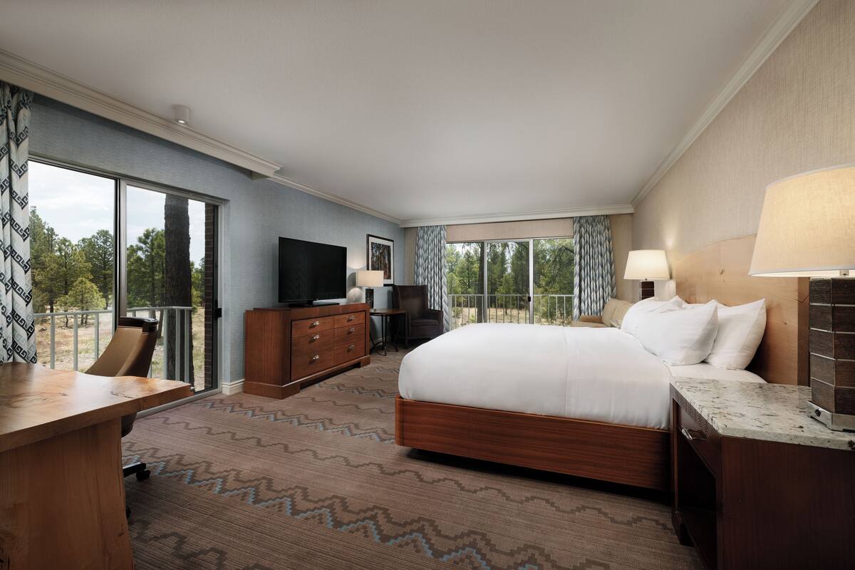 A hotel room with a large bed, a desk, a TV, and a balcony with a view of trees.