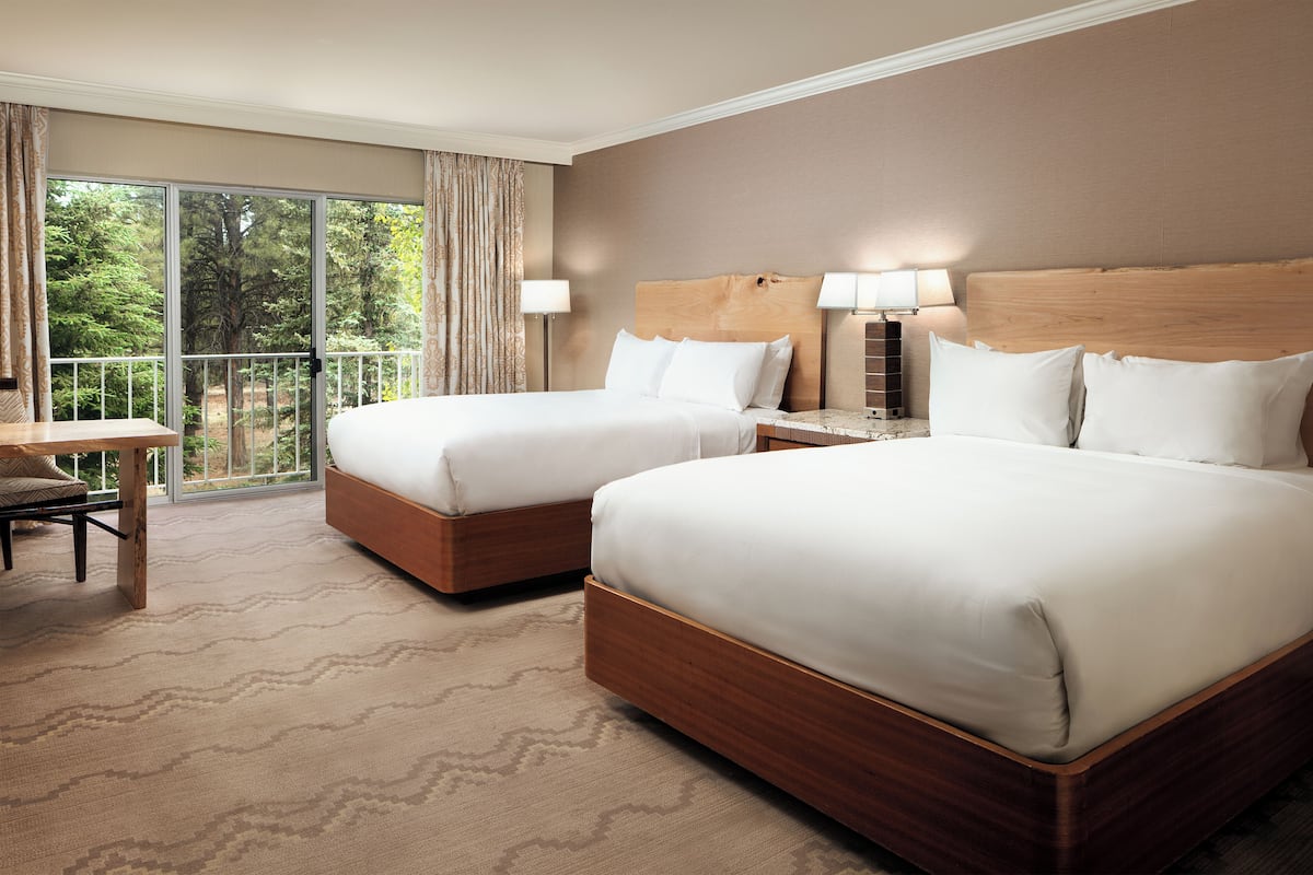 A hotel room with two beds, a desk, and a balcony with a view of trees.