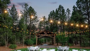 An outdoor wedding setup with round tables and chairs, a wooden archway, and string lights.