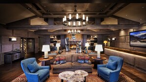 A spacious living room with a large sofa, two armchairs, a coffee table, and a chandelier. There are lamps on side tables, a fireplace, and a TV mounted on the wall.