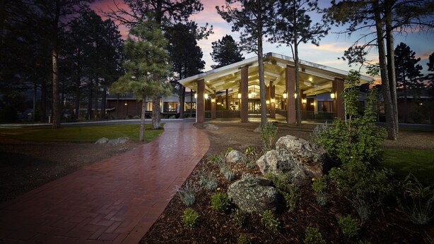 A modern building with a covered entrance, surrounded by trees and a landscaped area with rocks and plants.
