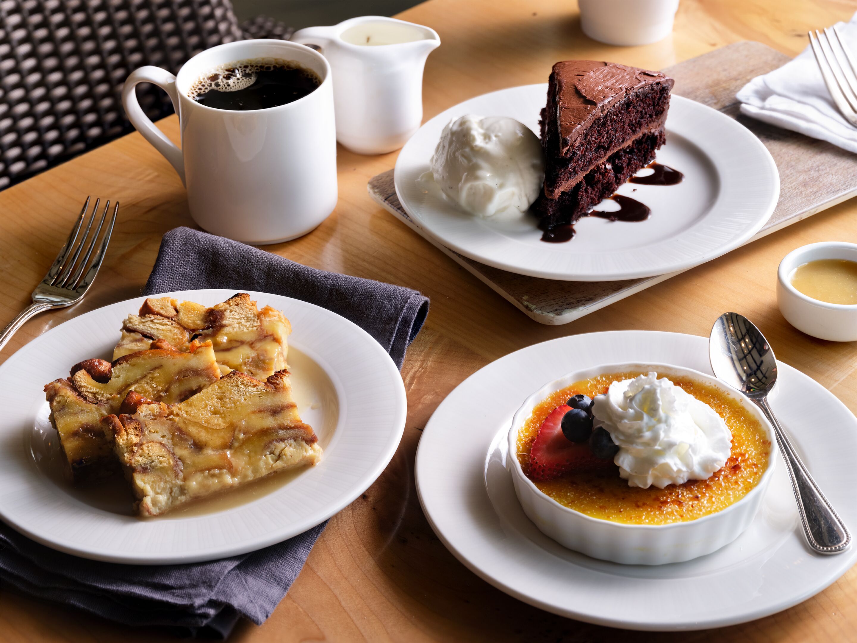 Uma mesa com uma fatia de torta, uma xícara de café, um pedaço de bolo com chantilly e uma tigela de sobremesa coberta com frutas e chantilly.