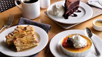 Uma mesa com uma fatia de torta, uma xícara de café, um pedaço de bolo com chantilly e uma tigela de sobremesa coberta com frutas e chantilly.