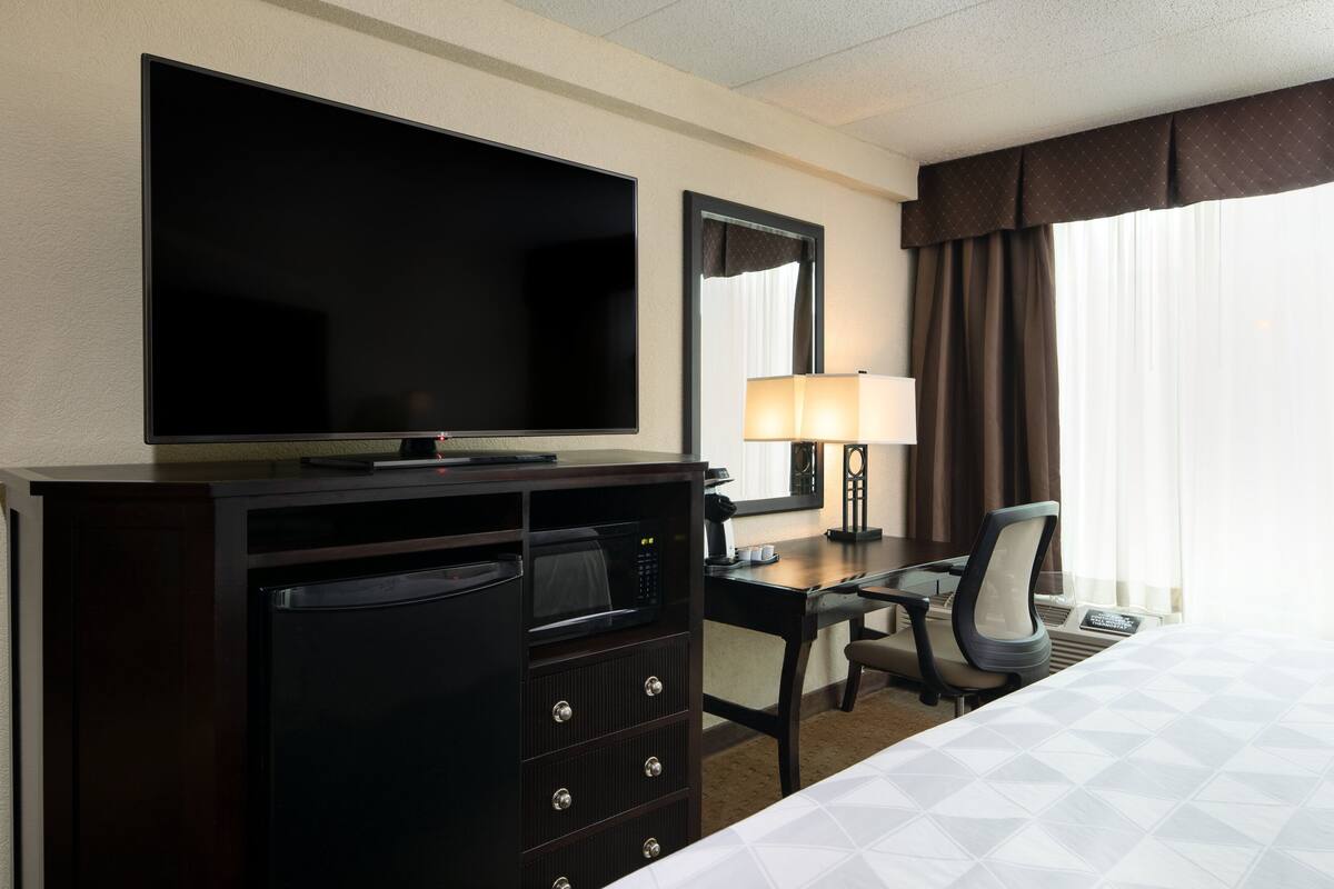 A hotel room with a bed, a desk with a lamp, a television, and a minibar.