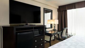 Quarto de hotel com uma cama, uma escrivaninha com luminária, televisão e frigobar.