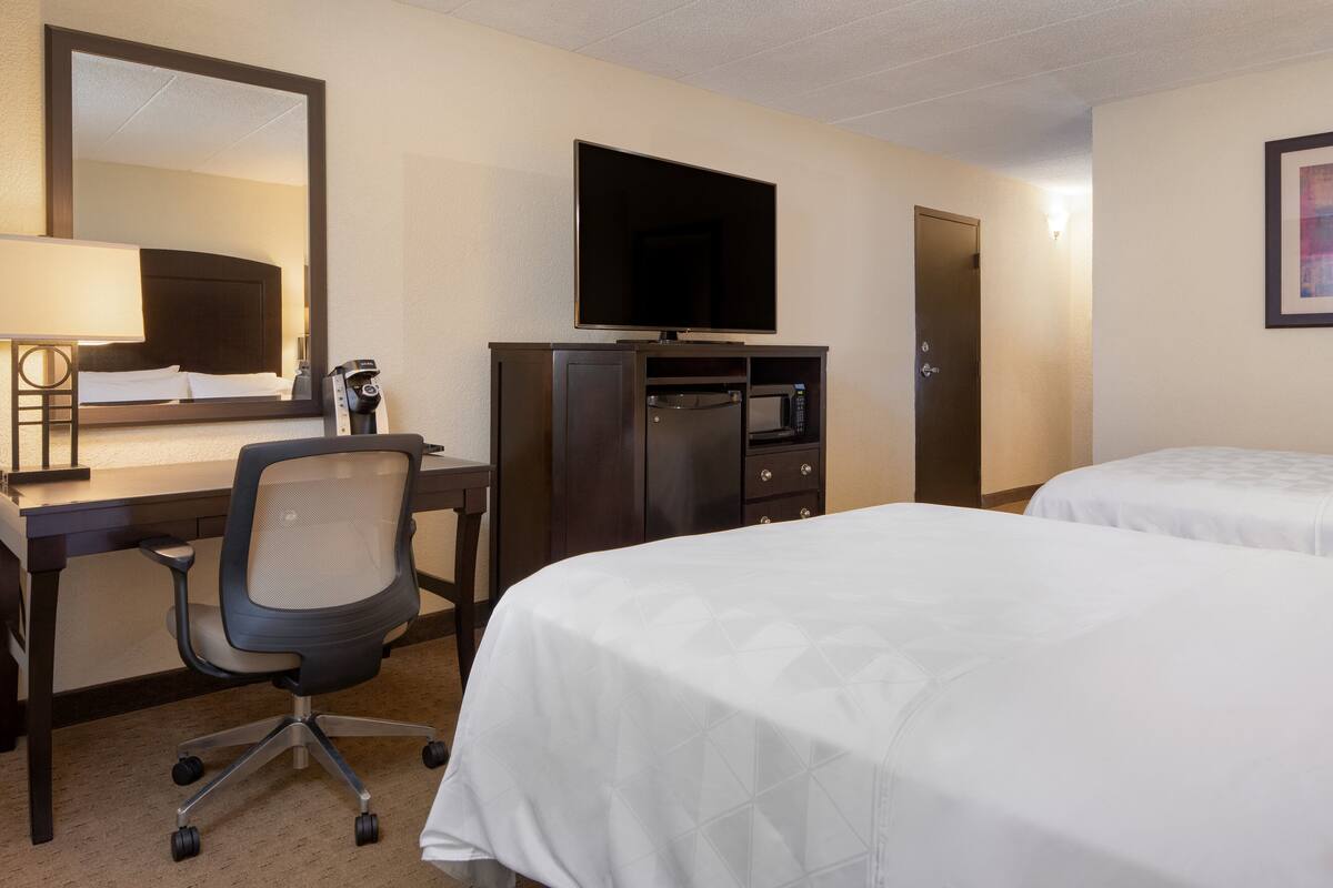 A hotel room with two beds, a desk with a chair, a television, a microwave, and a door.