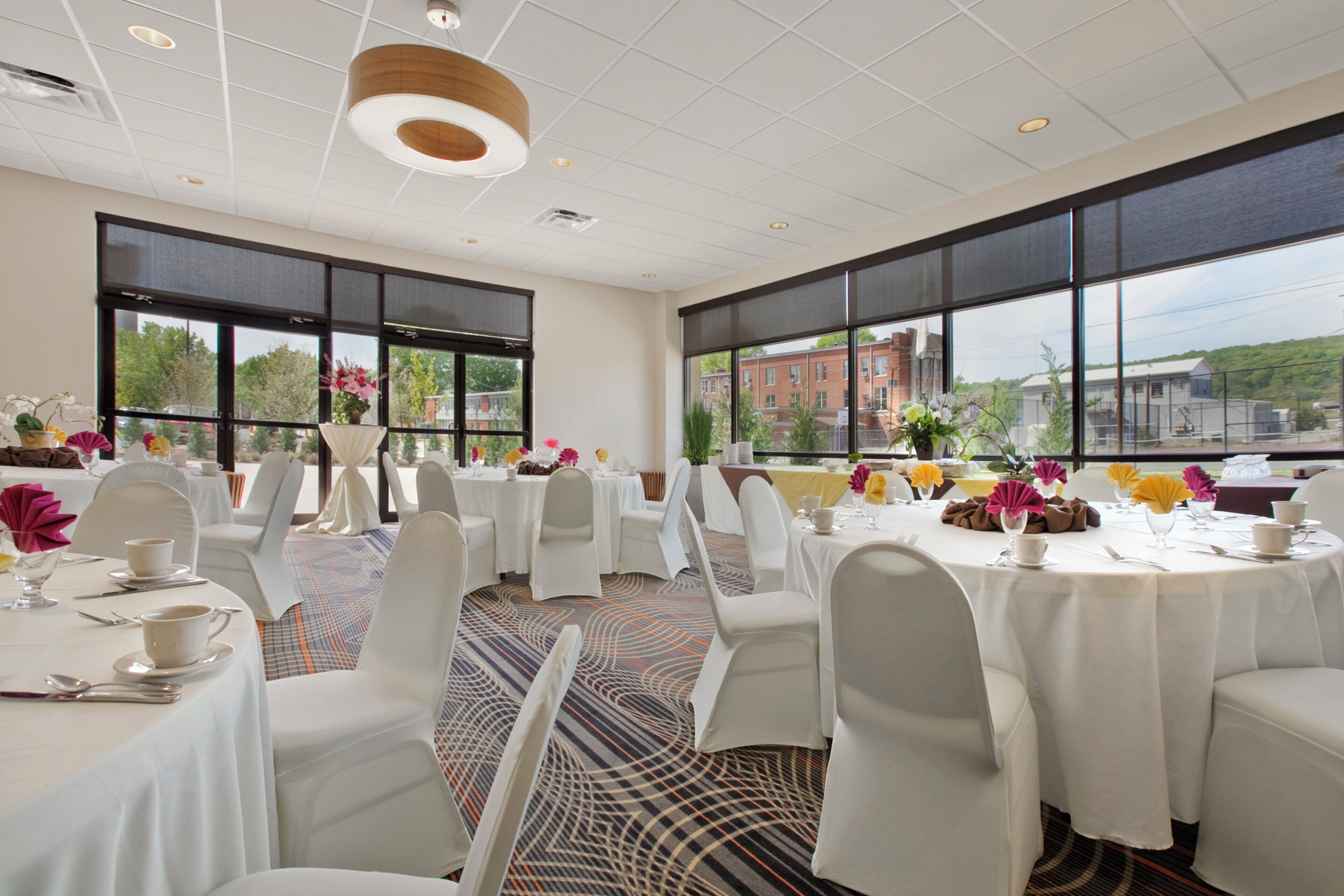 A conference room with round tables, white chairs, and large windows offering a view of buildings and trees.