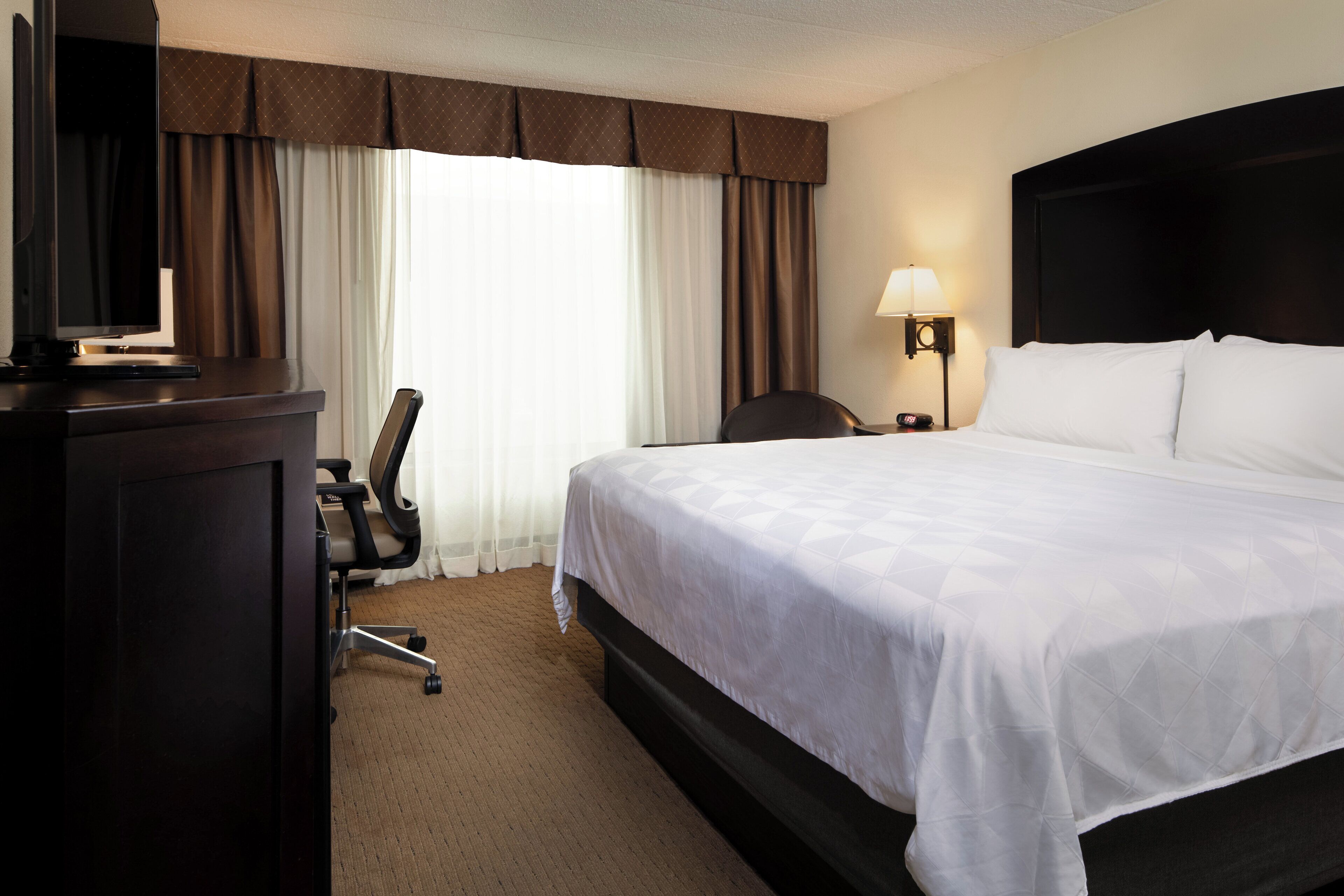 A hotel room with a bed, a desk with a computer, a chair, a lamp, and a window with curtains.