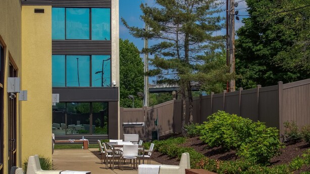 An outdoor patio area with white wicker furniture, a wooden fence, and a building with large windows. - Holiday Inn Hotel & Suites Charleston West by IHG (South Charleston)