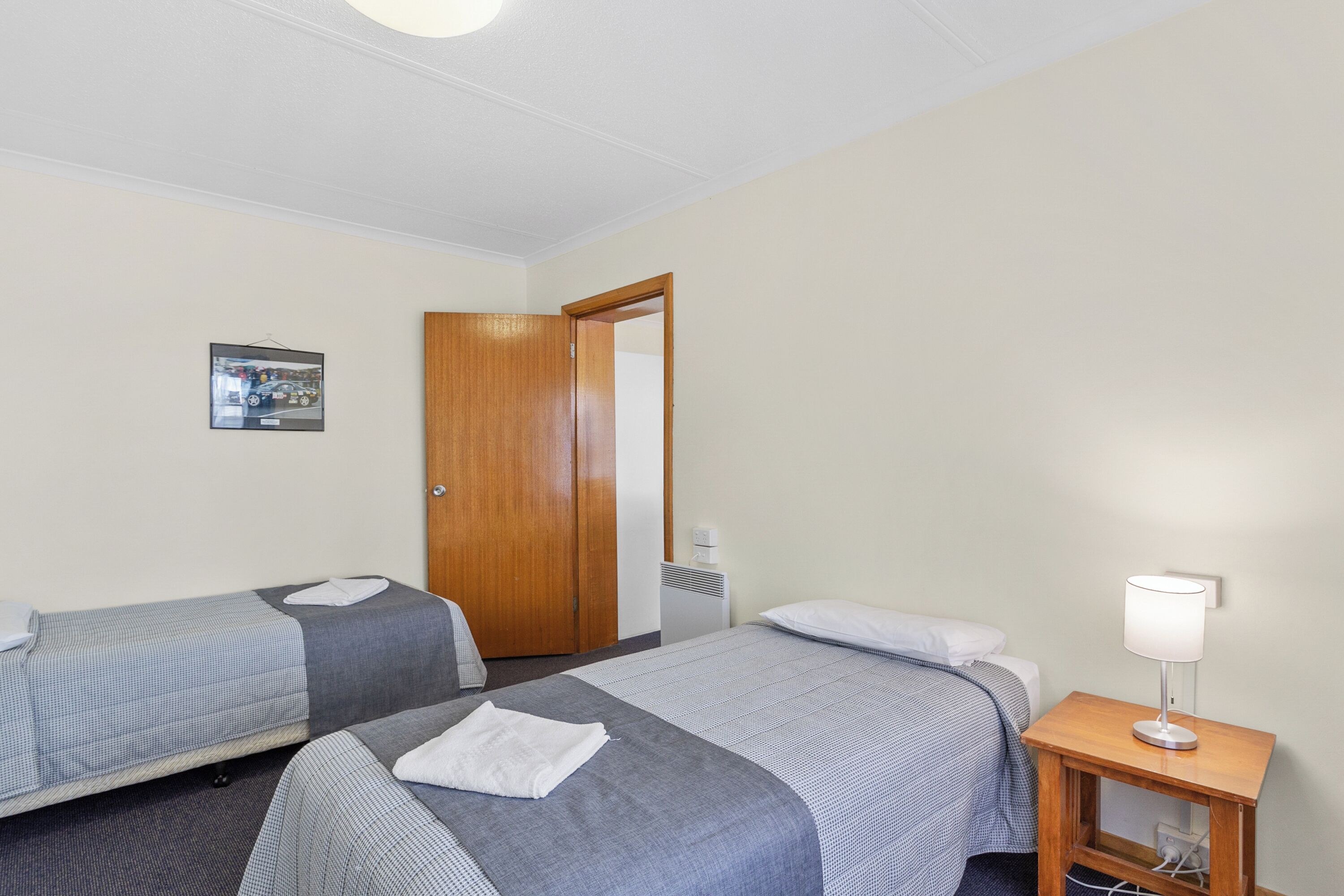 Habitación de hotel con dos camas, una mesita de noche de madera con una lámpara, una puerta y un cuadro enmarcado en la pared.