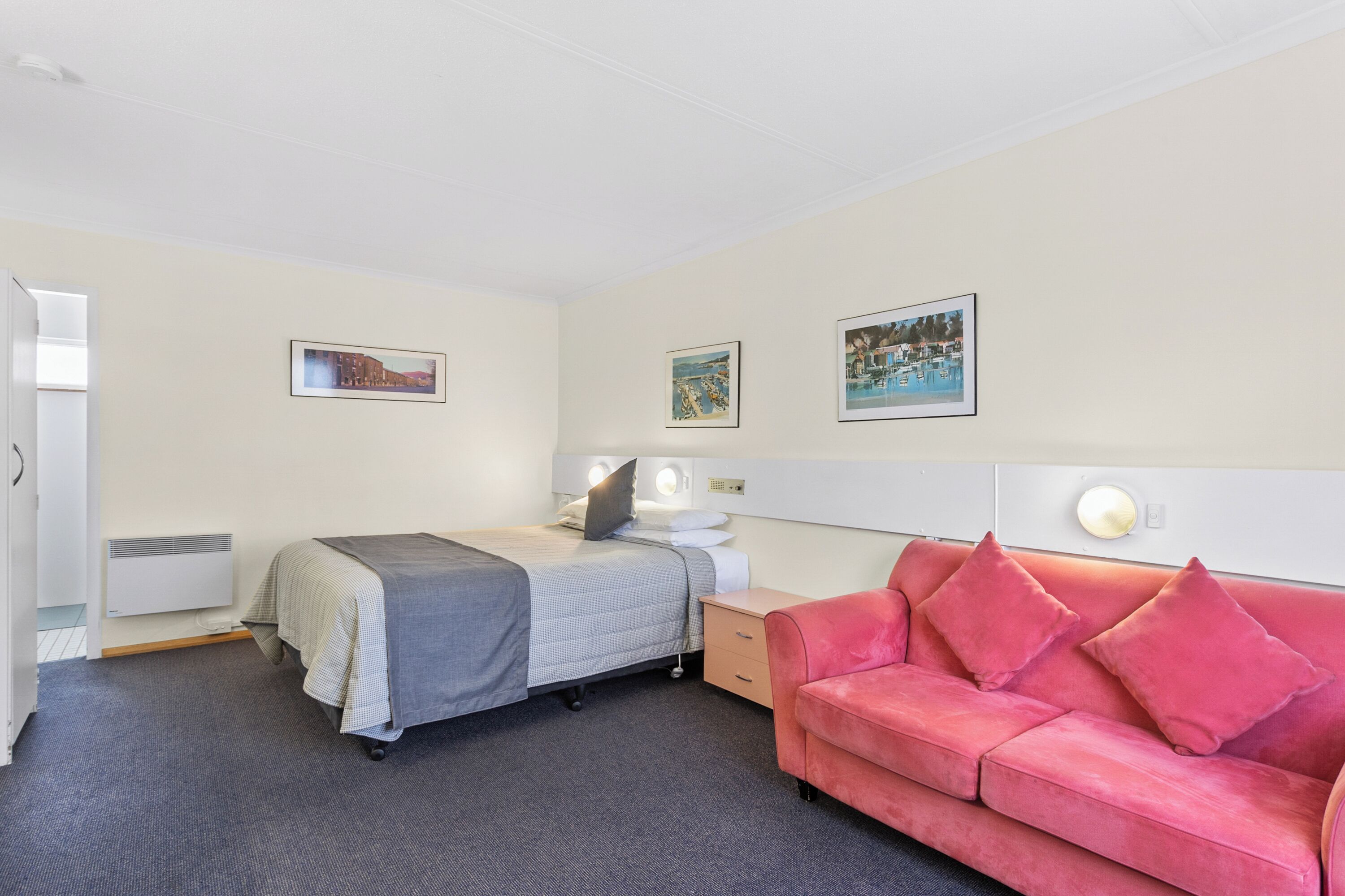 Une chambre d’hôtel avec un lit, un canapé rouge, une table de chevet avec une lampe et trois tableaux encadrés accrochés au mur.