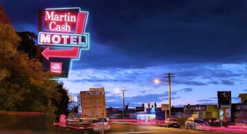 Un letrero de motel iluminado con neón, Martin Cash Motel, con vista despejada de la carretera y edificios circundantes.