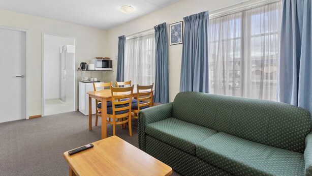 A room with a green sofa, wooden dining table, chairs, a kitchenette, and large windows with blue curtains. - Martin Cash Motel (Moonah)