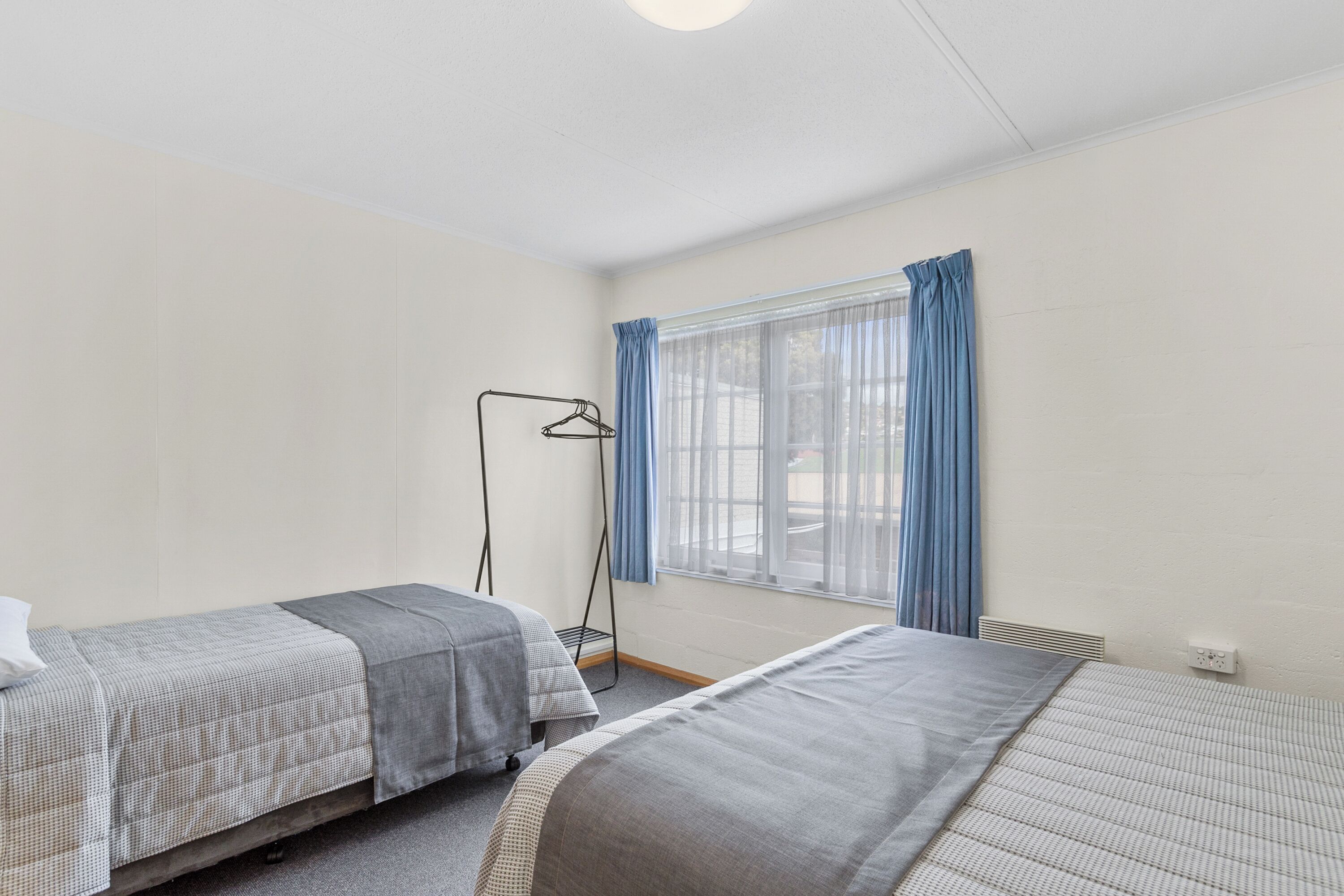 A room with two beds, a window with curtains, a floor lamp, and a radiator.