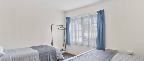 A room with two beds, a window with curtains, a floor lamp, and a radiator.