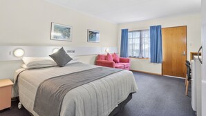 A hotel room with a bed, a pink chair, a wooden door, and a window with blue curtains. - Martin Cash Motel (Moonah)