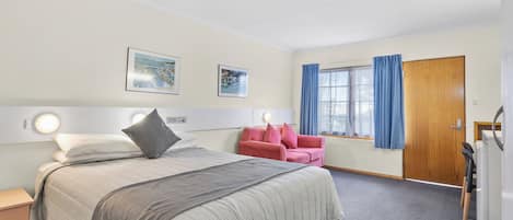 A hotel room with a bed, a pink chair, a wooden door, and a window with blue curtains.