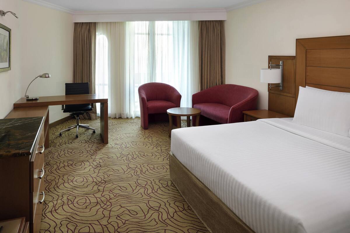 Ein Hotelzimmer mit Bett, Schreibtisch, zwei Stühlen, einem kleinen Tisch und einem Fenster mit Vorhängen.