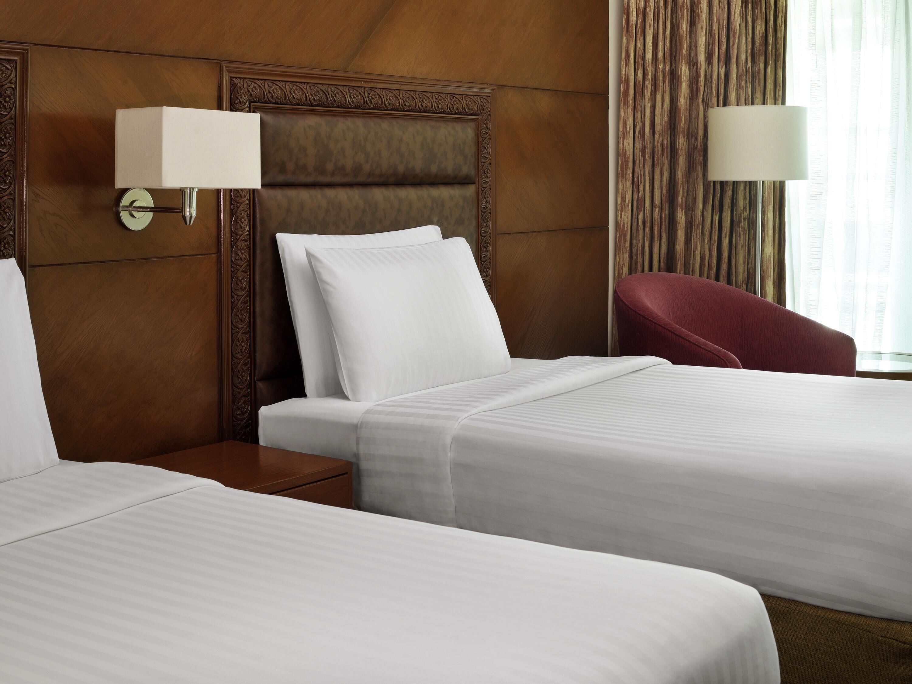 A hotel room with two beds, a nightstand, a red chair, and a lamp.